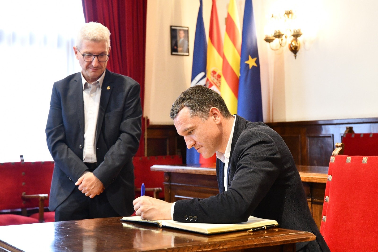 El conseller Oscard Ordeig sentado firmando el libro de honor en el salón de plenos del Ayuntamernt de Esplugues.Le acompaña el alcalde a su lado de pie.
